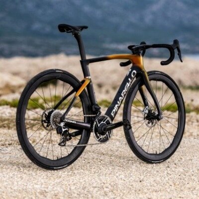 この画像は、Pinarello DOGMA Fの屋外撮影シーン。エアロ形状のカーボンフレームと洗練されたゴールド×ブラックのデザインが際立ち、最高峰のレーシングバイクとしての存在感を放っている。







